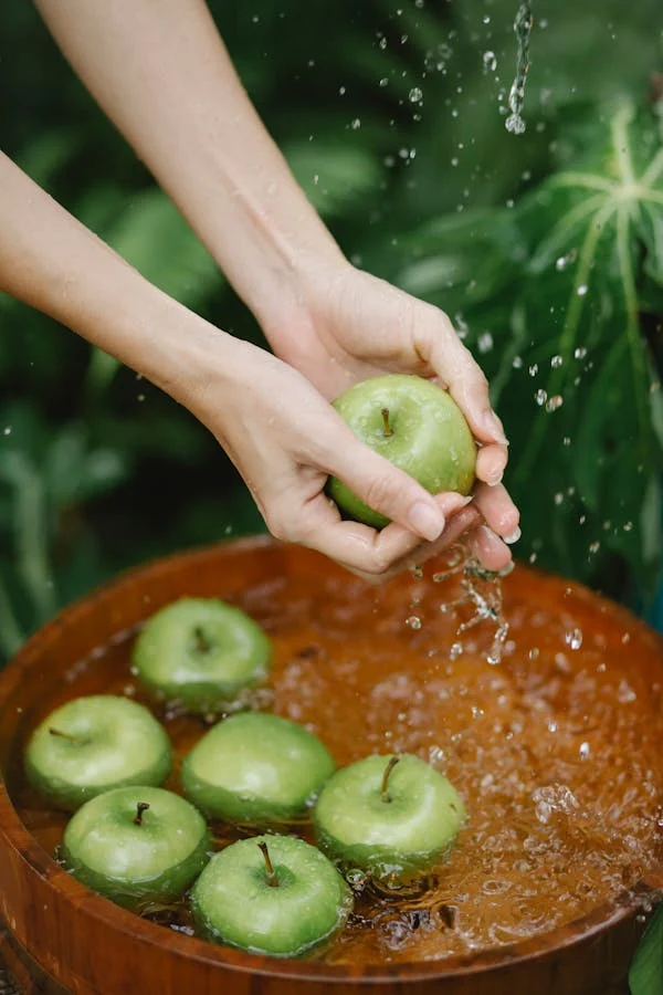 Hands holding a fresh green apple under splashing water over a wooden bowl filled with more green apples against a lush leafy background.